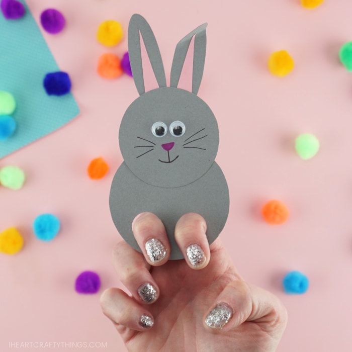 Close up image of a finished gray bunny finger puppet with an adult placing two finger in the holes, showing how to play with the puppet.